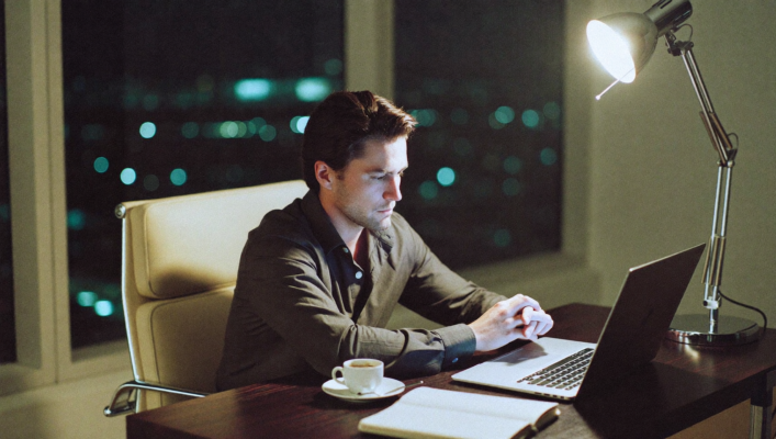 Hombre en su treinta y pocos sentado ante un escritorio minimalista moderno de noche, iluminado por una lámpara de diseño cálida, expresión muestra agotamiento pero concentración; taza de espresso vacía en el escritorio, portátil abierto frente a él, ventana grande a oscuras mostrando la ciudad de noche al fondo