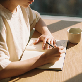 Manos escribiendo en un cuaderno de lino crudo sobre una mesa de roble claro, bajo la luz clara de la mañana que entra por una ventana, con una taza de cerámica mate junto al papel.
