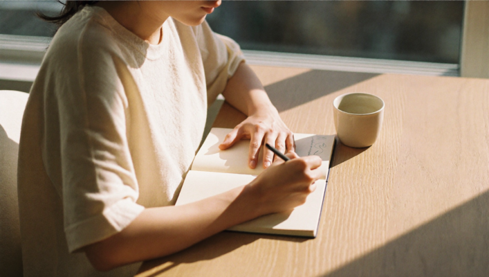 Manos escribiendo en un cuaderno de lino crudo sobre una mesa de roble claro, bajo la luz clara de la mañana que entra por una ventana, con una taza de cerámica mate junto al papel.