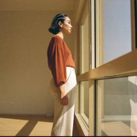 Una mujer joven de pie frente a un ventanal de piso a techo en un interior minimalista contemporáneo al amanecer, con suelo de roble claro y paredes crema, mirando hacia el exterior en calma, luz matinal clara proyectando sombras definidas en el espacio diáfano.