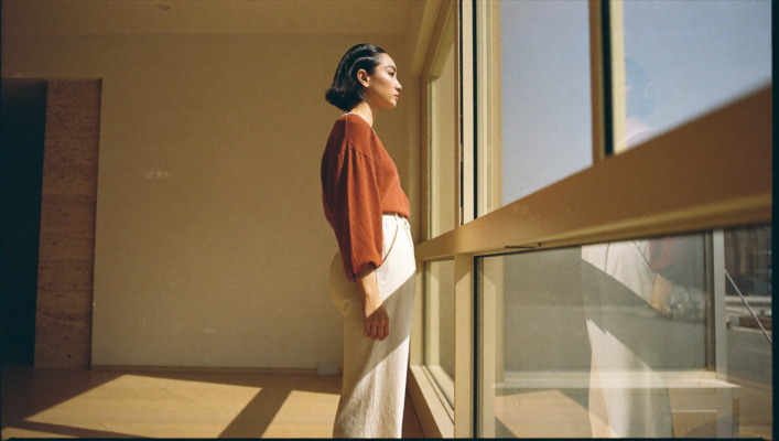 Una mujer joven de pie frente a un ventanal de piso a techo en un interior minimalista contemporáneo al amanecer, con suelo de roble claro y paredes crema, mirando hacia el exterior en calma, luz matinal clara proyectando sombras definidas en el espacio diáfano.