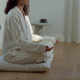 Persona meditando con los ojos cerrados y la espalda recta, sentada en un cojín de lino blanco en una habitación minimalista. Luz suave de amanecer filtra a través de cortinas de lino blancas, iluminando el suelo de roble pálido. Junto a ella descansa una taza de cerámica mate con té sobre travertino.