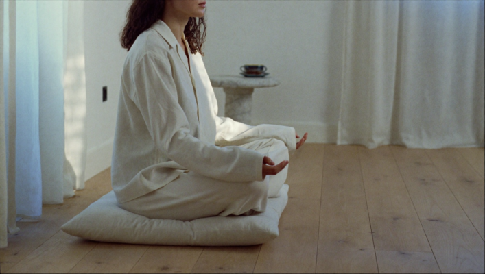 Persona meditando con los ojos cerrados y la espalda recta, sentada en un cojín de lino blanco en una habitación minimalista. Luz suave de amanecer filtra a través de cortinas de lino blancas, iluminando el suelo de roble pálido. Junto a ella descansa una taza de cerámica mate con té sobre travertino.
