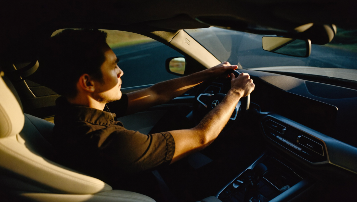 Persona en sus treinta años dentro de un auto de lujo minimalista, manos en el volante, mandíbula y cuello tensos pero rostro controlado, luz matinal dorada y azul cortando por parabrisas y ventanas laterales, carretera desenfocada adelante.