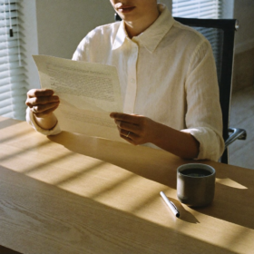 Una persona en una camisa de lino color crema se sienta en un escritorio de roble claro, leyendo una carta abierta. Sombras suaves de persianas venecianas cruzan la superficie de travertino. Una taza de café y una pluma descansan intactas a un lado. La luz de la tarde filtra suavemente por la ventana.