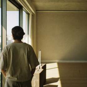 Una persona en sus treinta años de pie junto a una gran ventana que da a una ciudad nueva, observando dos maletas de viaje en el piso de madera clara de un apartamento vacío bañado por luz matutina.