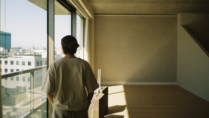 Una persona en sus treinta años de pie junto a una gran ventana que da a una ciudad nueva, observando dos maletas de viaje en el piso de madera clara de un apartamento vacío bañado por luz matutina.
